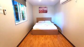 A bedroom with a wooden bed, a window, an air conditioner, and a cityscape painting on the wall.
