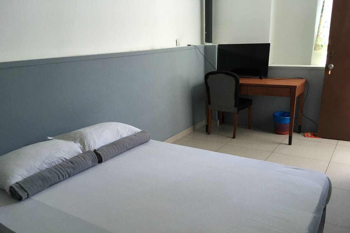 Ein Einzelbett mit weißer Bettwäsche, einem grauen Kissen und einer grauen Decke. Ein Holztisch mit Stuhl und Fernseher sind vorhanden. Eine blaue Mülltonne ist in der Ecke sichtbar.