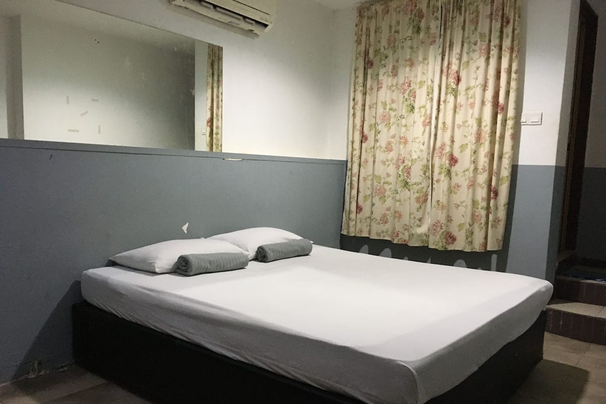 Ein Einzelbett mit grauer Matratze und Kissen, ein Fenster mit blumigem Vorhang, ein Spiegel über dem Bett und ein Lichtschalter an der Wand.