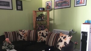 A living room with a black sofa, floral cushions, a wooden shelf with decorative items, a white table with a vase, and a refrigerator. - Ideal Hotel Sri Permaisuri Cheras (Kuala Lumpur)
