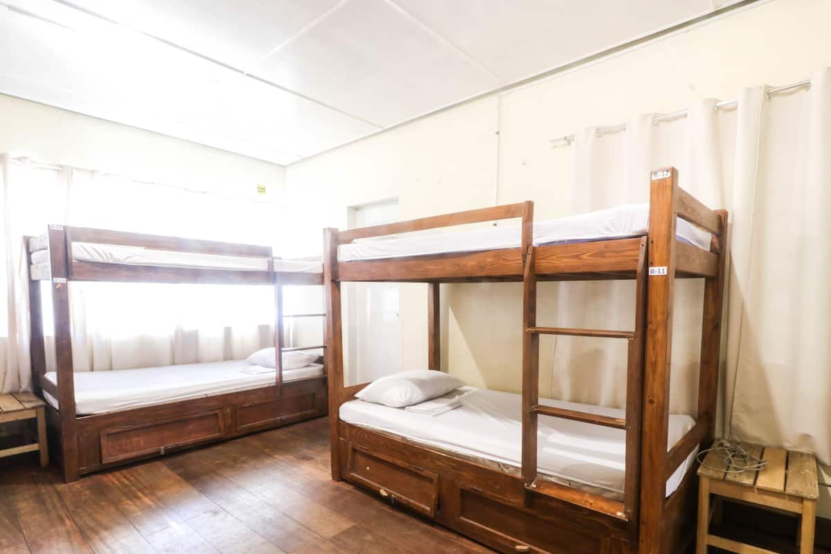 Quarto com duas beliches, piso de madeira e cortinas brancas.