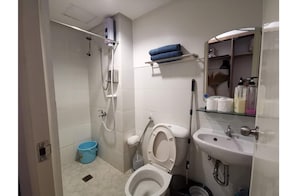A bathroom with a toilet, sink, and shower. There is a blue bucket and a shelf with towels. - SweetSuites at Pine Crest New Manila (Quezon City)