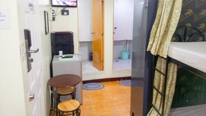 A compact kitchen with a refrigerator, a small table with stools, a television mounted on the wall, and a bathroom visible through an open door. - 8 Hostel at Quiapo (Manila)