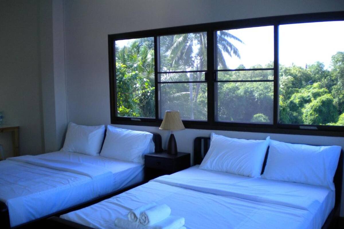 A room with two beds, a nightstand with a lamp, and a window with a view of greenery.