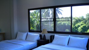 A room with two beds, a nightstand with a lamp, and a window with a view of greenery. - Sripiamsuk (Pathum Thani)