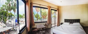 A hotel room with a bed, a small table, and a chair. There are large windows with curtains, and a view of palm trees and a street outside. - G.I Lodge by Zuzu (Pattaya)