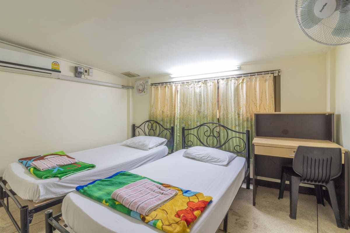 A room with two single beds, a desk, a chair, and a standing fan.