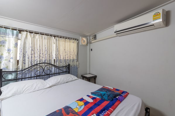 A single bed with a white sheet, a colorful blanket, and a headboard. There is a small bedside table with a lamp, and an air conditioning unit mounted on the wall.