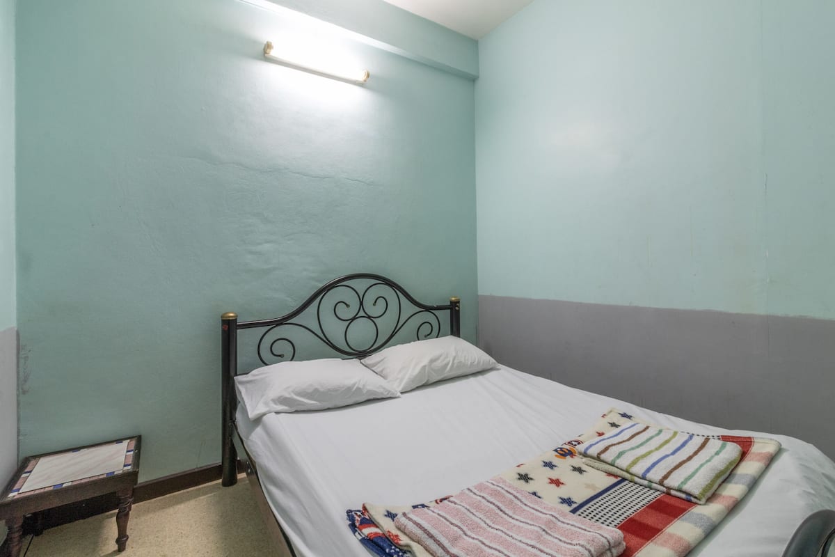 A single bed with a metal frame, white bedding, and a colorful blanket. There is a small wooden table beside the bed.