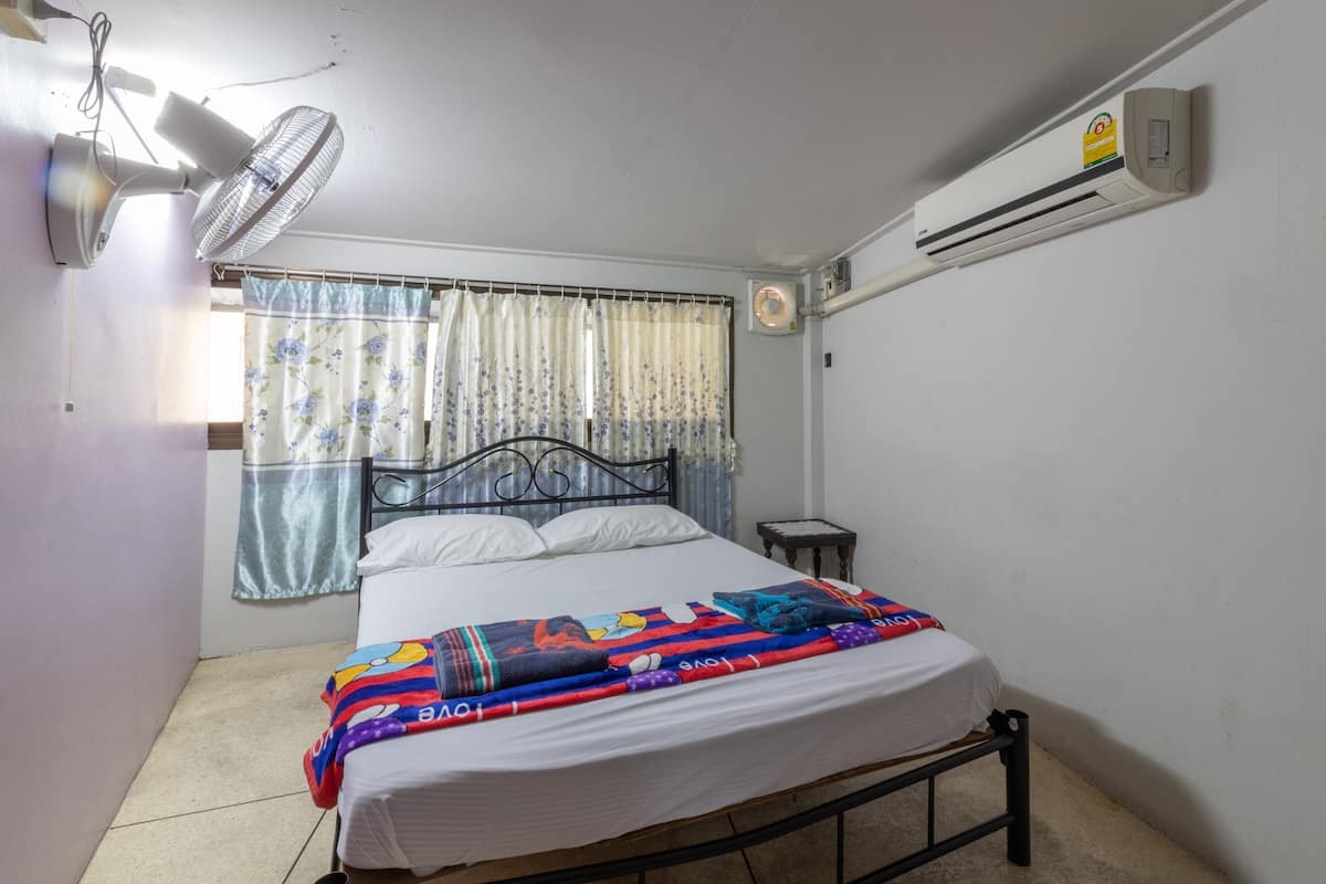 A bedroom with a single bed, a standing fan, an air conditioner, and a window with floral curtains.