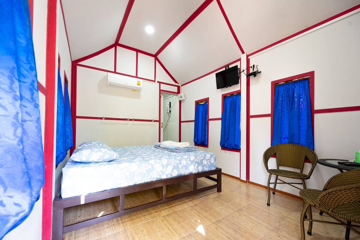 Quarto com cama, cadeira, mesa pequena, televisão e janela com cortinas.