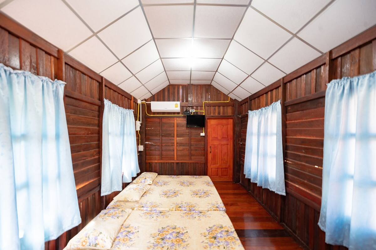 Quarto com paredes de madeira, teto azulejado, cama com roupa de cama floral e cortinas nas janelas.