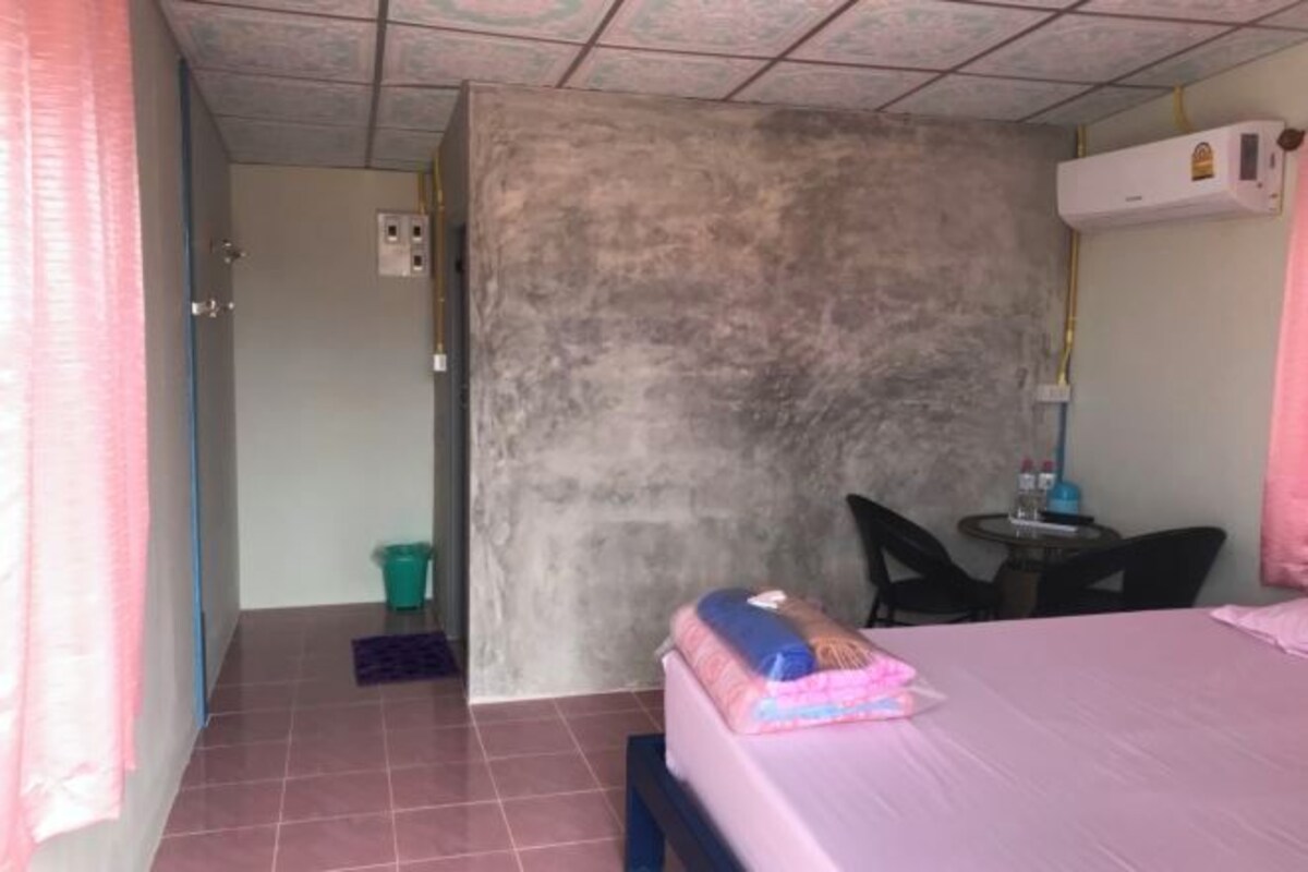 Una habitación pequeña y funcional con cama, escritorio y silla. Hay un cubo de basura y una mesita con lámpara.
