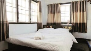 A neatly made bed in a room with large windows and curtains, a wooden headboard, and a small bedside table with a lamp. - Center Home City (Chiang Mai)