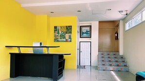 A reception area with a desk, a yellow wall, a framed cityscape painting, and a staircase leading to an upper level. - Andaman Place (Thep Krasattri)