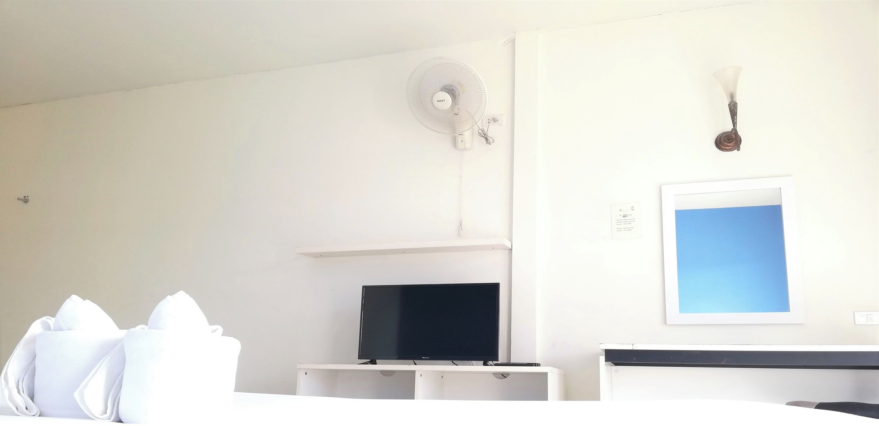 A hotel room with a bed, a TV on a stand, a wall-mounted fan, and a light fixture.
