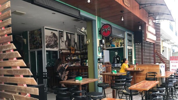 An outdoor café with wooden tables and black stools, a bar area, and a staircase leading to an upper level. - JT Guesthouse (Kamala)