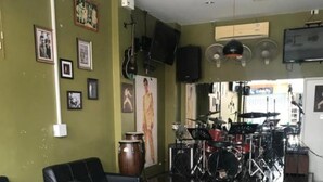 A modern lounge area with black leather sofas, a small wooden table, and a drum set in the background. - JT Guesthouse (Kamala)