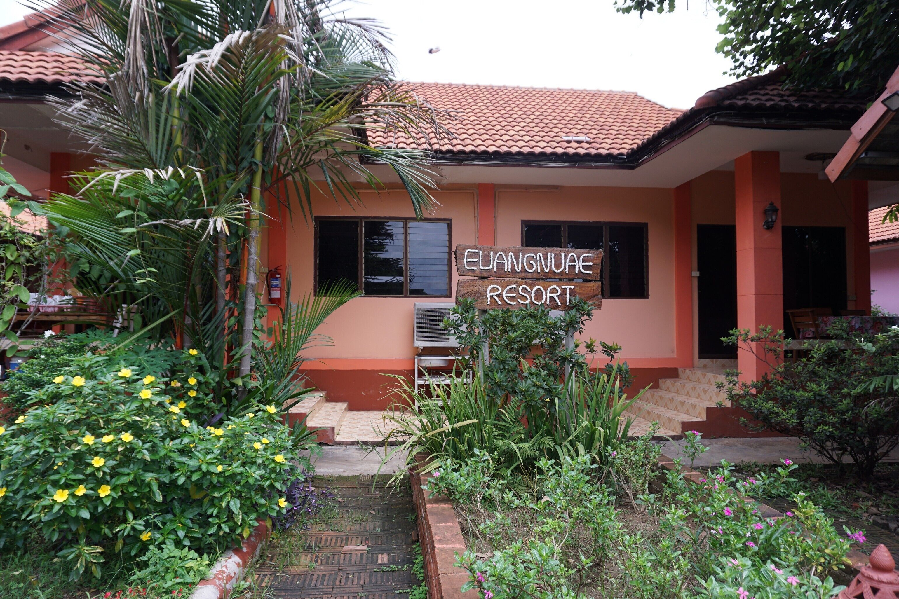 A building with a sign reading "Euangnuae Resort." It has a tiled roof, a staircase leading to the entrance, and is surrounded by various plants and flowers.