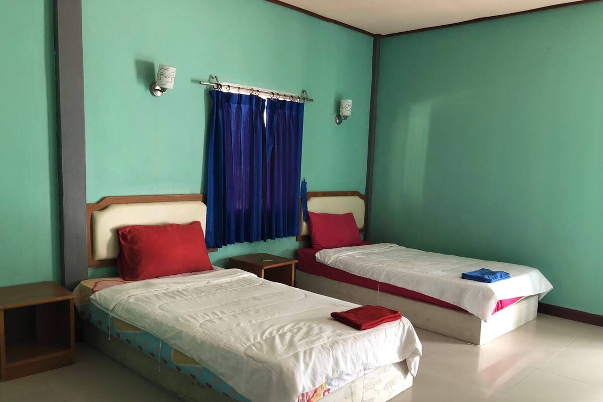 A hotel room with two single beds, each with a red pillow and a white bedspread. There are two nightstands, one with a lamp on each side of the beds. The room has teal walls and a window with blue curtains.