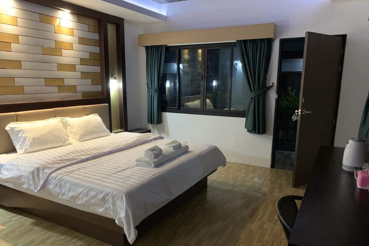 A modern hotel room with a large bed, a wooden headboard, a bedside table with a kettle, and a view of the outdoors through a window with curtains.