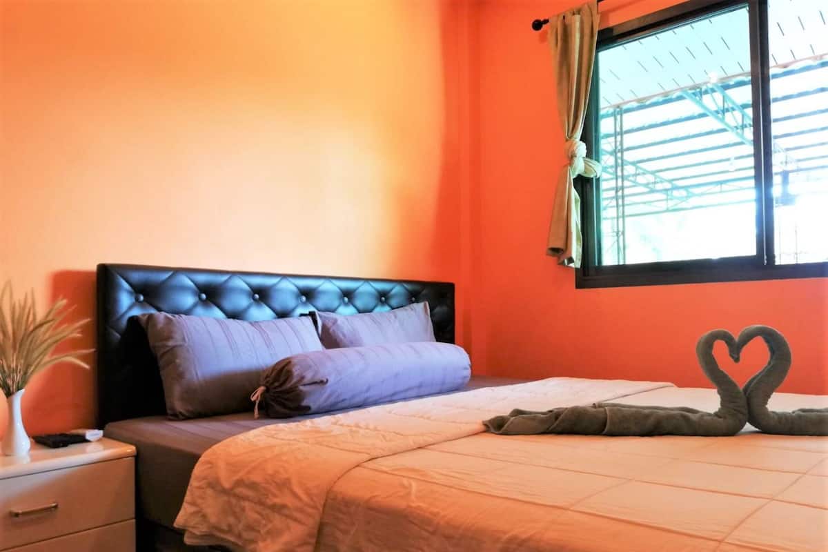 Une chambre à coucher avec une tête de lit capitonnée, un lit agrémenté d’un arrangement de coussins en forme de cœur, une table de chevet avec une lampe et une fenêtre avec des rideaux.