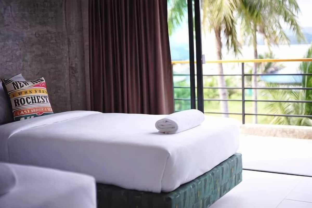 Una cama con colchón blanco y almohada con texto, cabecera verde y vista a palmeras a través de una ventana.