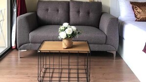 Una sala de estar con un sofá gris, una mesa de centro con un jarrón de flores y un cuadro de un sendero en el bosque en la pared.