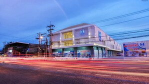 A modern building with multiple storefronts, including a visible 'M' logo and a sign indicating 'OPEN NOW!'. - Manonta Budget Hotel (Hat Yai)