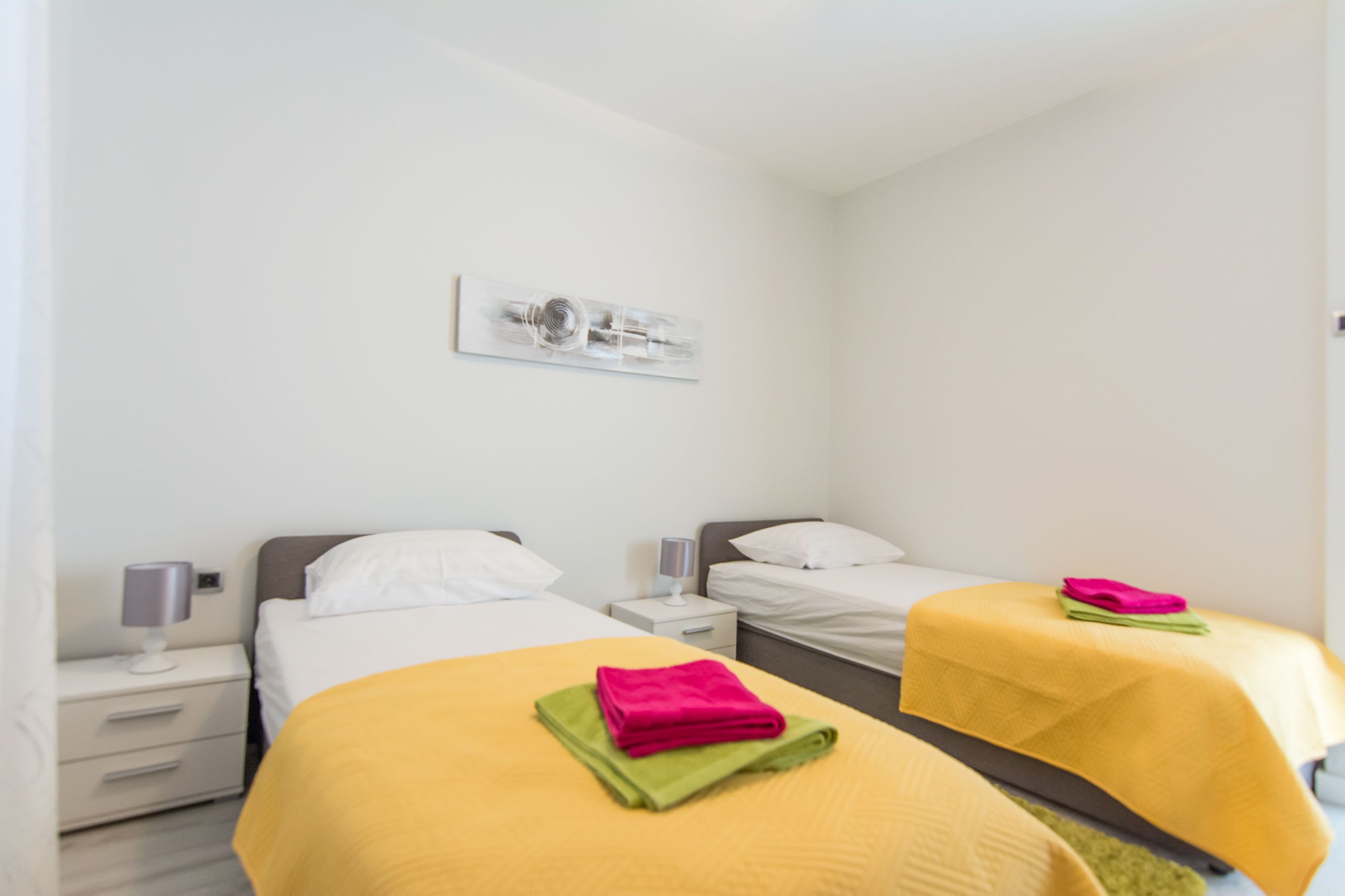 Habitación de hotel con dos camas, cada una con una colcha amarilla y una toalla doblada. Hay una mesita de noche con una lámpara y una pequeña mesa con otra lámpara. Las paredes son blancas, y hay un cuadro enmarcado en la pared.