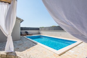 A rectangular swimming pool with clear blue water, surrounded by stone tiles and a white curtain. - House Moonstone (Zadar)