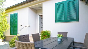 Un patio al aire libre con una mesa de madera oscura y sillas de mimbre, rodeado de plantas en macetas y una pared blanca con contraventanas verdes.