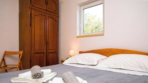 Una cama bien hecha con ropa de cama blanca y dos toallas enrolladas, un armario de madera, una silla y una ventana con luz diurna.