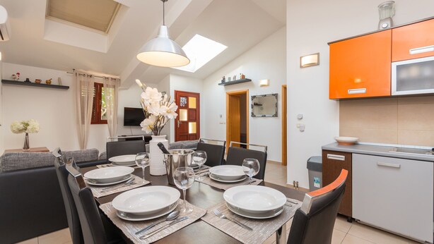Uma sala de jantar moderna com mesa posta para quatro, uma cozinha com armários laranja e uma sala de estar com sofá e televisão.