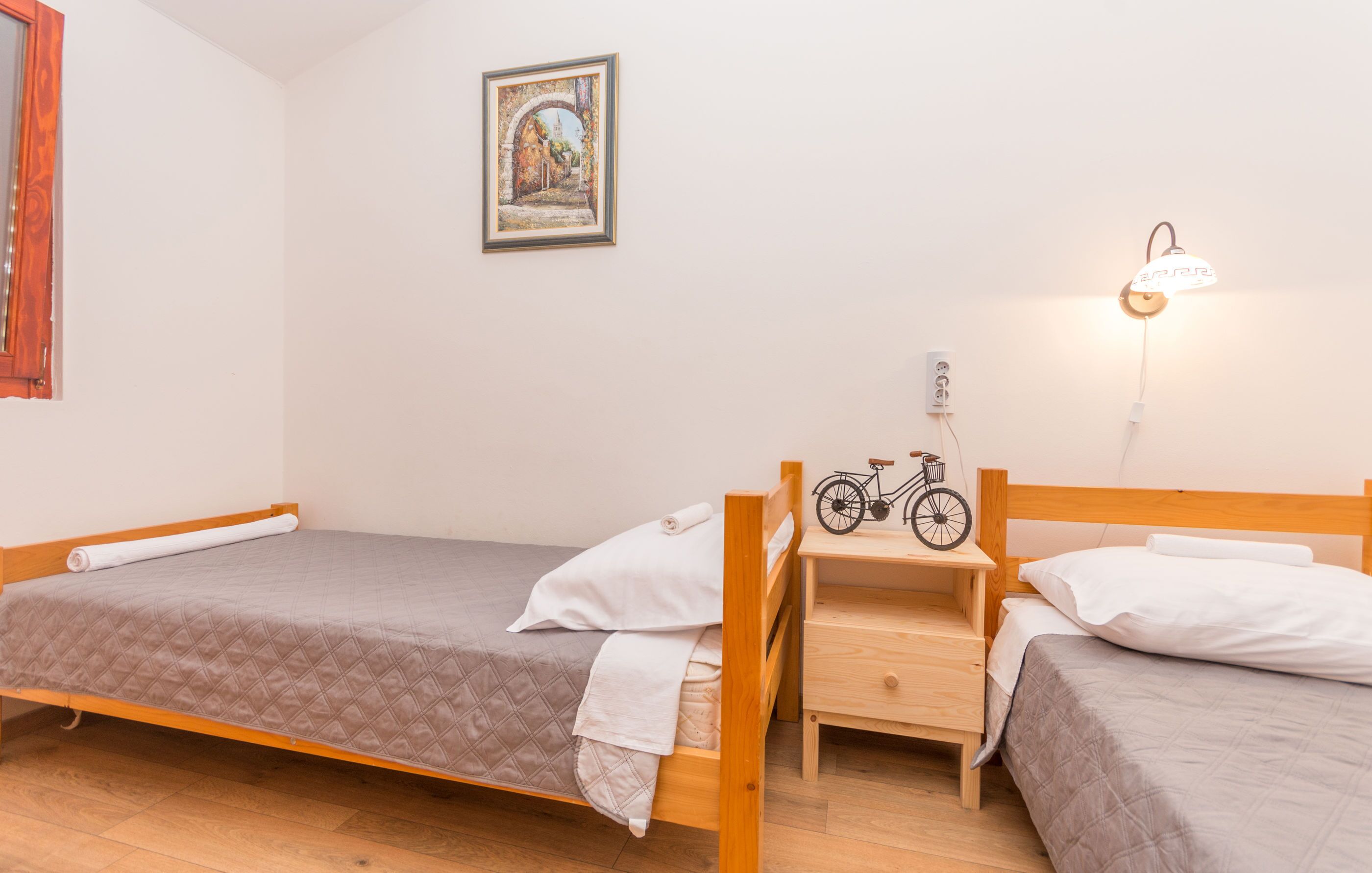 A small, clean room with two single beds, a wooden nightstand, a bicycle, and a wall-mounted lamp.