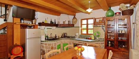 A kitchen with a wooden ceiling, a refrigerator, a dining table with a bowl of fruit, and a cabinet with dishes.