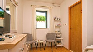A compact room with a wooden floor, a TV, a small table with two chairs, a window with curtains, a wooden door, and a shelf with various items. - Apartment Bilberry (Biograd)