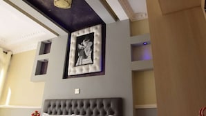 Een kamer met een gestoffeerd hoofdeinde, een ingelijst schilderij aan de muur, en een plafond met een klok en inbouwverlichting.