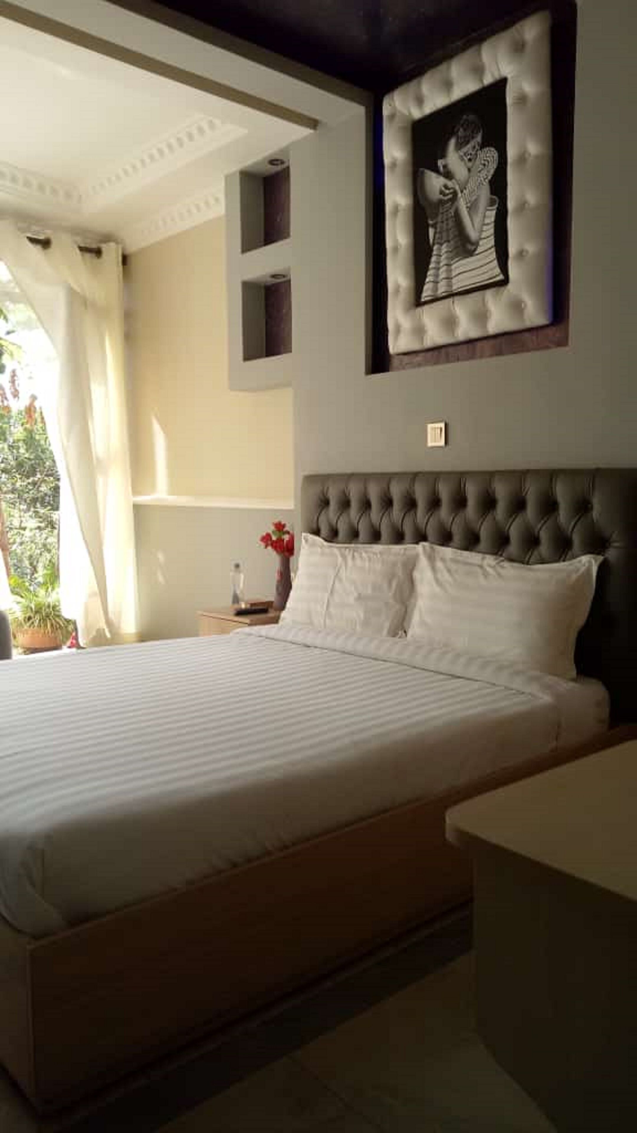A neatly made bed with white linens in a room with a tufted headboard, a framed picture on the wall, and a small bedside table with a vase of flowers.