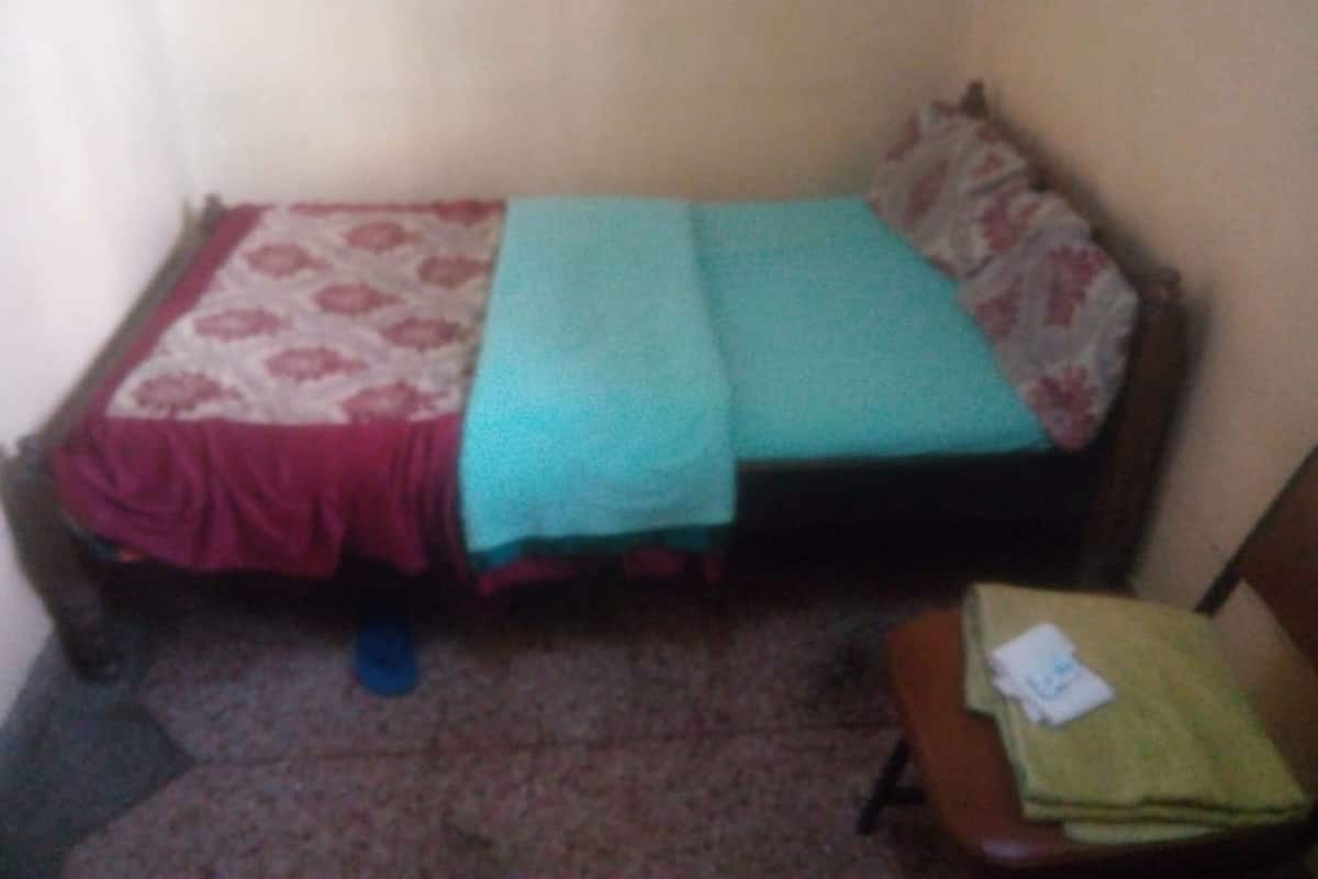 A small, single-bed room with a patterned bedspread, a teal blanket, and a green pillow. There is a chair with a green cushion and a white item on it. The floor is carpeted, and the walls are plain.
