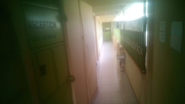 A long, narrow hallway with a reception area on the left, tiled flooring, and a green metal railing on the right.