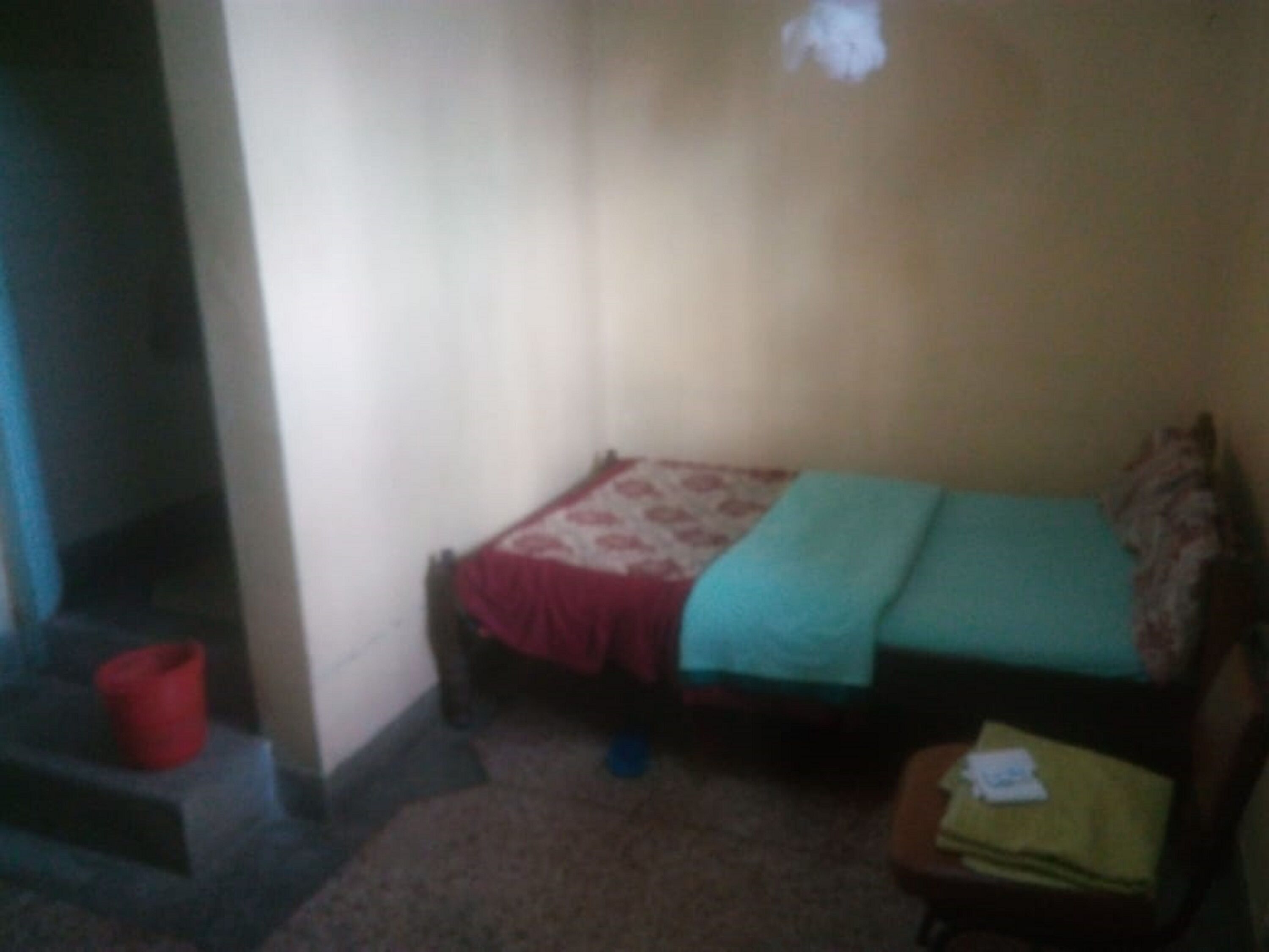 A small, dimly lit room with a single bed, a small table, and a red bucket.