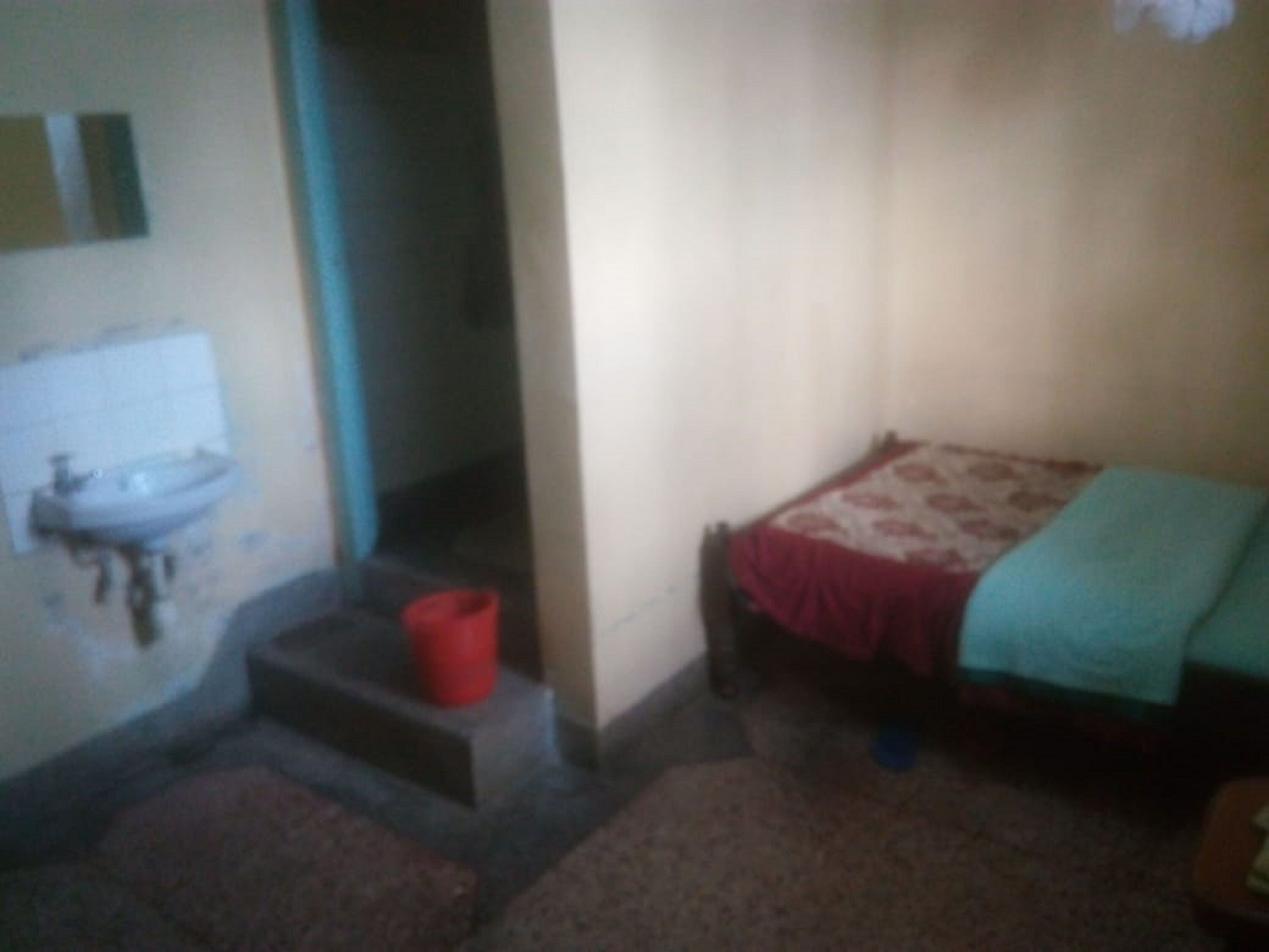 A small, dimly lit room with a single bed, a red bucket, and a sink against the wall.