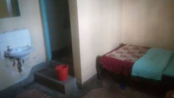 A small, dimly lit room with a single bed, a red bucket, and a sink against the wall.