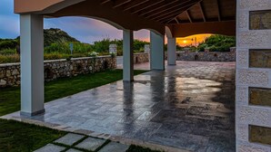 Un espace extérieur couvert, aménagé en patio, avec des murs en pierre et un sol carrelé, offrant une vue sur un coucher de soleil en arrière-plan.