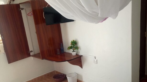 A bedroom with a bed, a bedside table with a plant, a hanging light, and a wooden door. - Marra Hill Hotel (Nakuru)