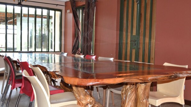 A dining area with a large wooden table surrounded by chairs, large windows, and a door with vertical stripes. - Olerai Hotel (Kitengela)