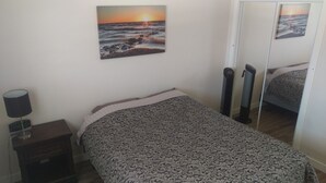 Quarto com colcha estampada, abajur, relógio digital e pintura de um pôr do sol sobre o oceano.