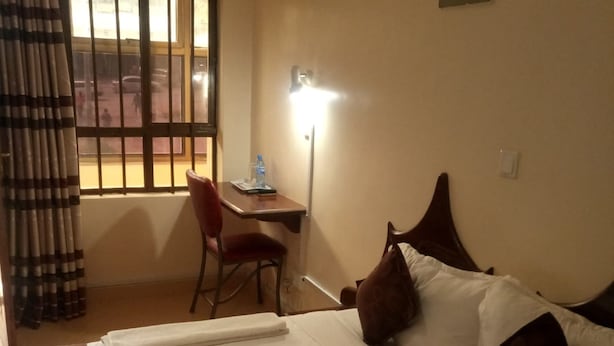 A hotel room with a bed, a desk, a chair, a nightstand, and a window with bars. - Remiz Guest House (Nakuru)