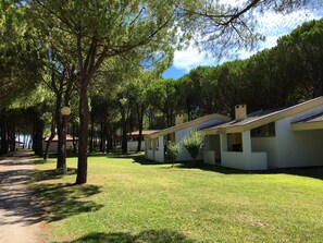 A row of modern, single-story buildings with a flat roof, surrounded by a well-maintained lawn and tall trees. - Club Del Sole Marina Julia Family Collection (Monfalcone)